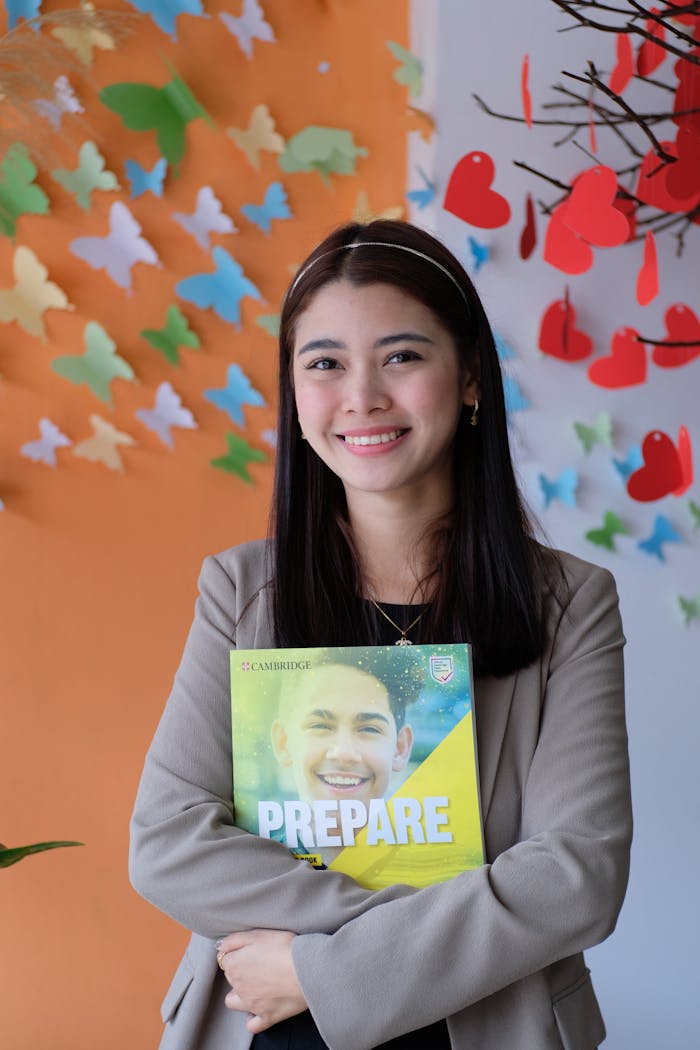 Smiling young woman holding a Cambridge book in a vibrant educational environment.